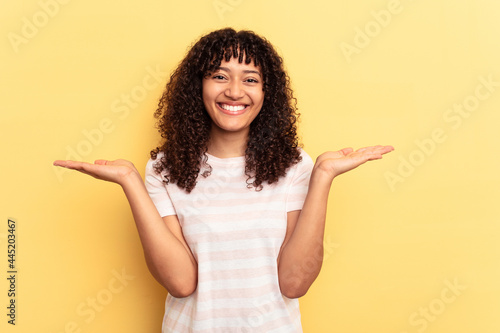 Young mixed race woman isolated on yellow background makes scale with arms, feels happy and confident.