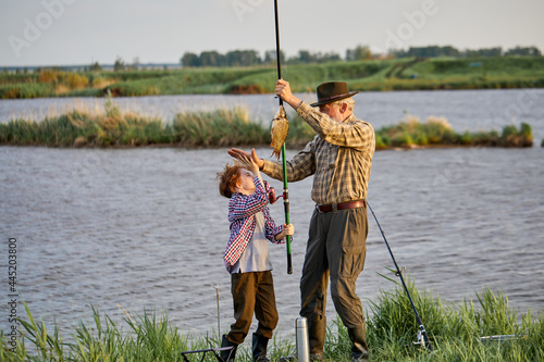 Summertime image of skilled fisherman on retirement having rest with grandson in wild nature using fishing rod, happy to catch fish, smile, give high five. Family, leisure and active hobby photo