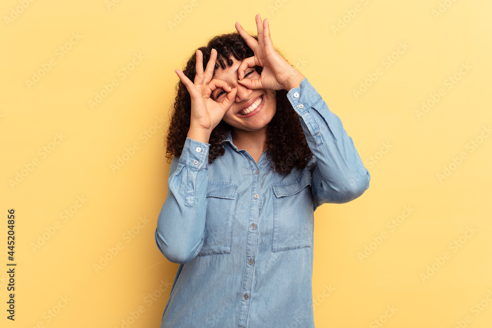 Young mixed race woman isolated on yellow background showing okay sign over eyes Young mixed race woman isolated on yellow background showing okay sign over eyes