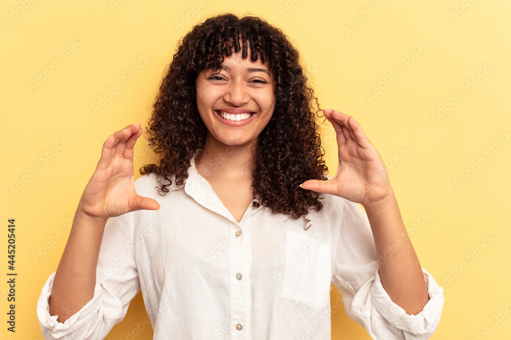 Young mixed race woman isolated on yellow background holding something with palms, offering to camera. Young mixed race woman isolated on yellow background holding something with palms, offering to camera.