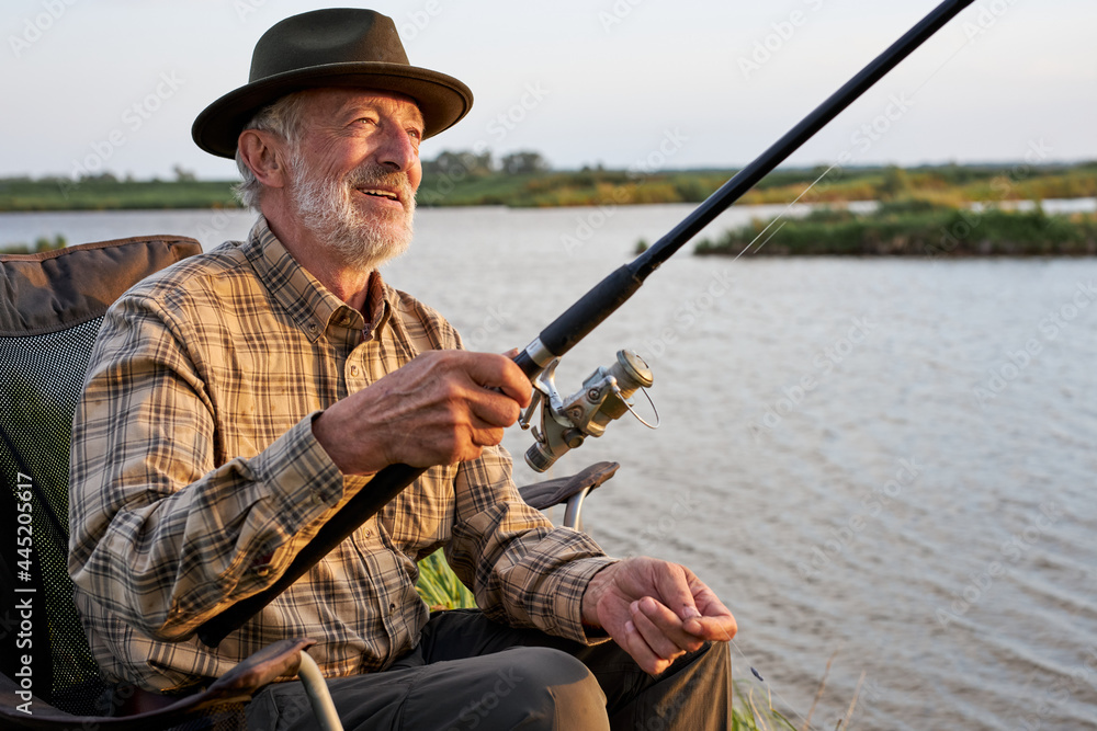 Elderly Nice Caucasian Man On Camping Holiday With Fishing Rod, Gra-haired Sit On Chair Alone Outdoors In Countryside Nature. Relax Leisure Time Elderly Nice Caucasian Man On Camping Holiday With Fishing Rod, Gra-haired Sit On Chair Alone Outdoors In Countryside Nature. Relax Leisure Time