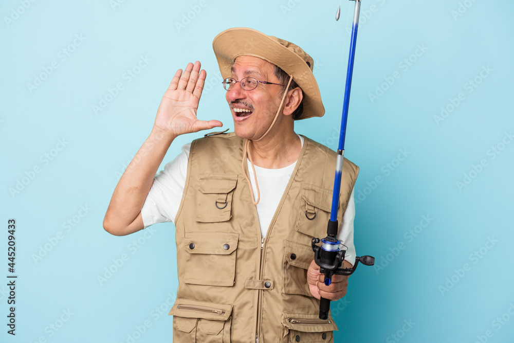 Senior indian fisherman holding rod isolated on blue background shouting and holding palm near opened mouth. Senior indian fisherman holding rod isolated on blue background shouting and holding palm near opened mouth.