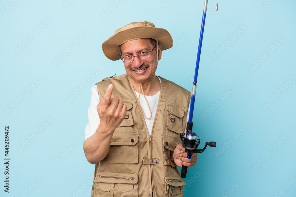 Senior indian fisherman holding rod isolated on blue background pointing with finger at you as if inviting come closer. Senior indian fisherman holding rod isolated on blue background pointing with finger at you as if inviting come closer.