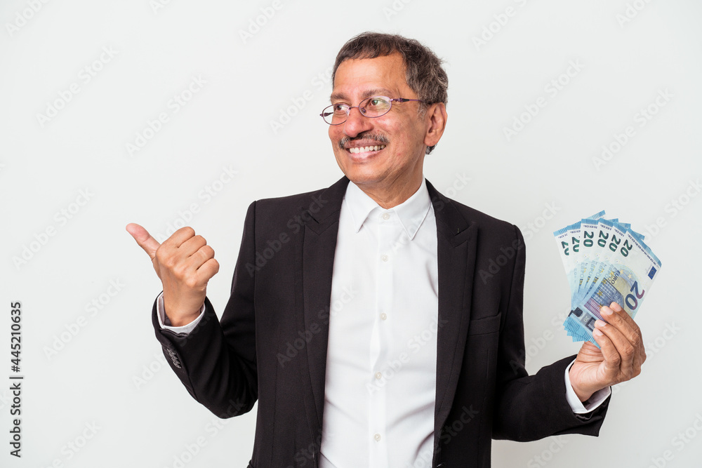 Middle aged indian business man holding bills isolated on white background points with thumb finger away, laughing and carefree. Middle aged indian business man holding bills isolated on white background points with thumb finger away, laughing and carefree.