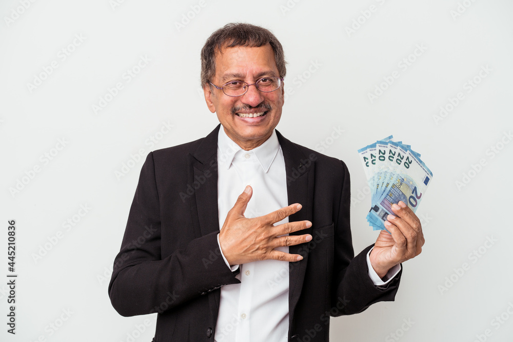 Middle aged indian business man holding bills isolated on white background laughs out loudly keeping hand on chest. Middle aged indian business man holding bills isolated on white background laughs out loudly keeping hand on chest.