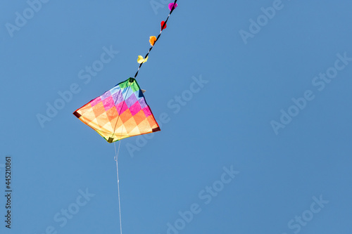 Child's rainbow coloured diamond kite with tail flying high in clear blue summer sky