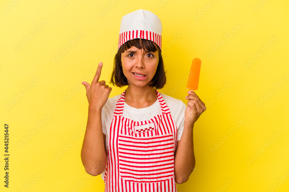 Young mixed race ice cream maker woman holding an ice cream isolated on yellow background showing a disappointment gesture with forefinger.