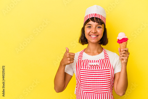 Young mixed race ice cream maker woman holding an ice cream isolated on yellow background smiling and raising thumb up © Asier
