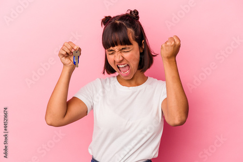 Young mixed race woman holding a keys isolated on pink background raising fist after a victory, winner concept.