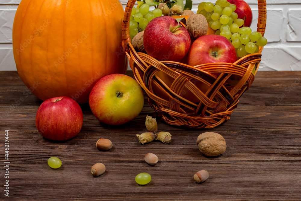 Orange pumpkins with apples and pears in a wicker basket with grapes, nuts and leaves on a brown wooden table against a white brick wall background. Close-up. Autumn still life. Orange pumpkins with apples and pears in a wicker basket with grapes, nuts and leaves on a brown wooden table against a white brick wall background. Close-up. Autumn still life.