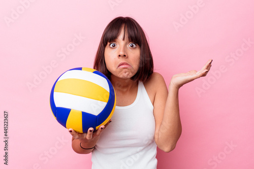 Young mixed race woman playing volleyball isolated on pink background shrugs shoulders and open eyes confused.