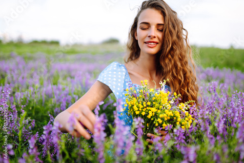 Young woman in stylish summer dress feeling free in the field with flowers in sunshine. Nature, vacation, relax and lifestyle. Summer landscape.