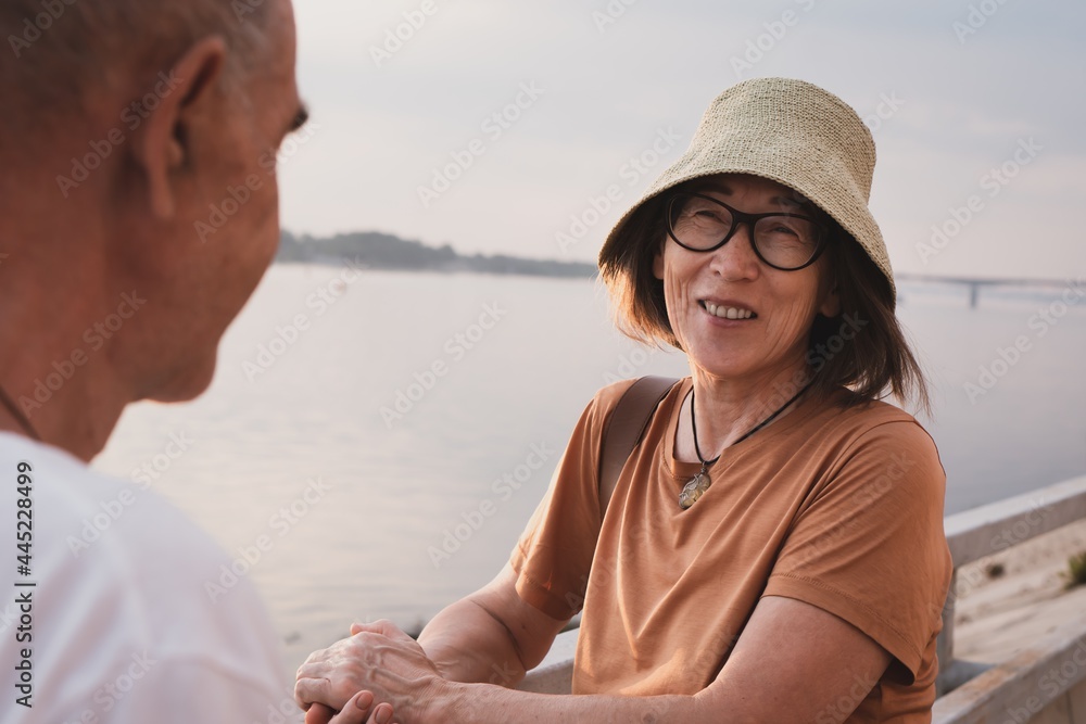smiling pretty Asian senior woman in hat and glasses with husband on sea or river branch at summer evening 