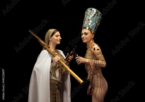 Foto Young beautiful woman in image of Nefertiti in art performance with her servant drinking beer isolated on dark background