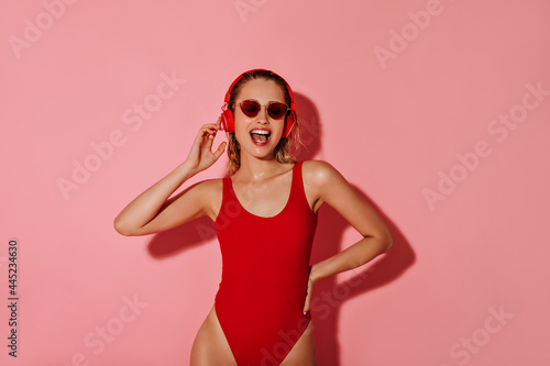 Positive girl with headphone in modern red sunglasses and bright swimming suit listening to music on pink isolated background..