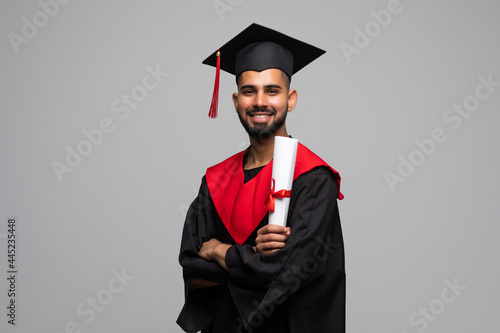 education, graduation and people concept - happy smiling indian male graduate student in mortar board and bachelor gown with diploma over grey background photo