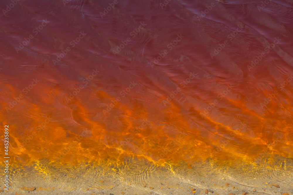 Textured surface of red poisoned muddy lake water near sandy shore in shallow water. There are small ripples on the water, there are reflections of light. Water pollution, background, copy space.