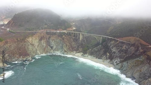 Cloudy aerial view of Bixby Bridge in Big Sur State Park in California. Vegetation and ocean waves can be seen along the coastline on Highway 1.