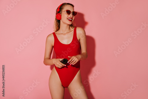 Happy woman with wet hair in sunglasses, headphone and red trendy swimming suit, smiling and holding bottle with soda water on pink backdrop..