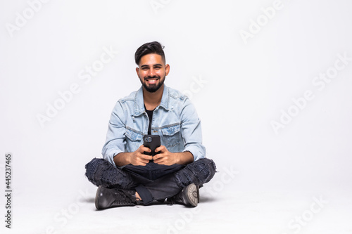 Full length of a cheerful young man sitting with legs crossed isolated over white background, using mobile phone