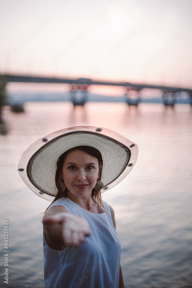 a young, happy woman in a straw hat stretches out her hand with an open palm to the camera and invites you to go on a summer vacation or travel to the river bank with a bridge at sunset. 