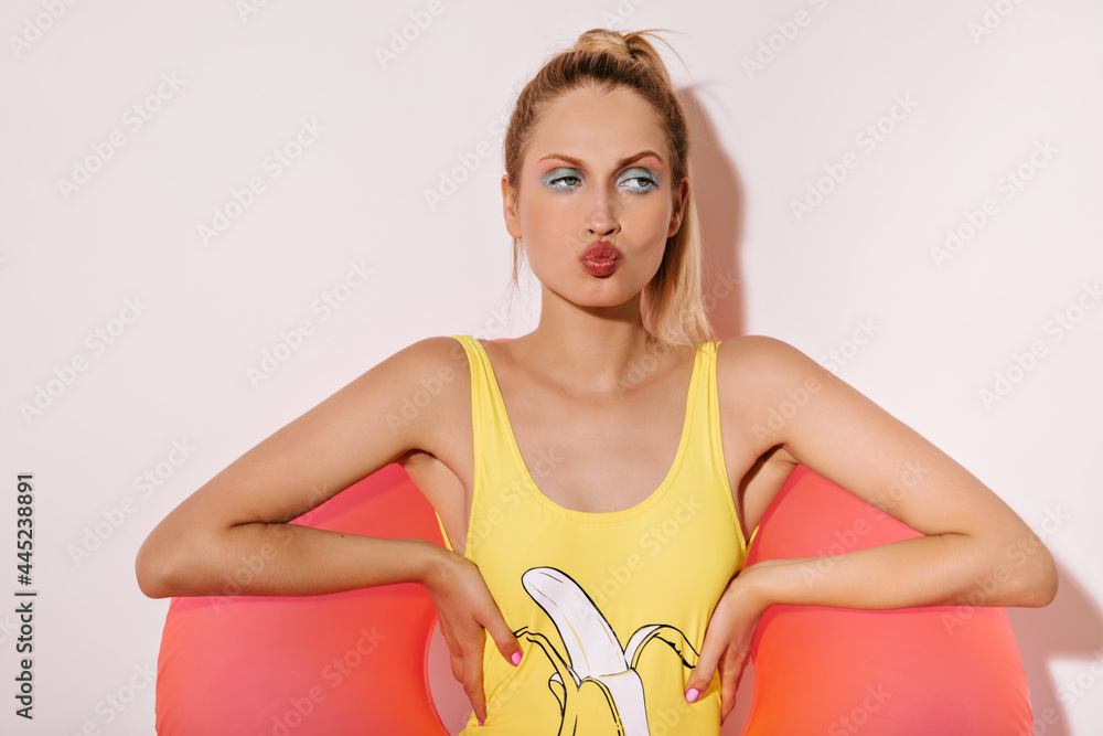 Cool blonde haired girl with trendy makeup in yellow swimsuit and posing with large pink swimming ring on isolated white background.. Cool blonde haired girl with trendy makeup in yellow swimsuit and posing with large pink swimming ring on isolated white background..