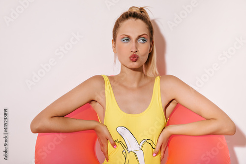 Cool blonde haired girl with trendy makeup in yellow swimsuit and posing with large pink swimming ring on isolated white background..
