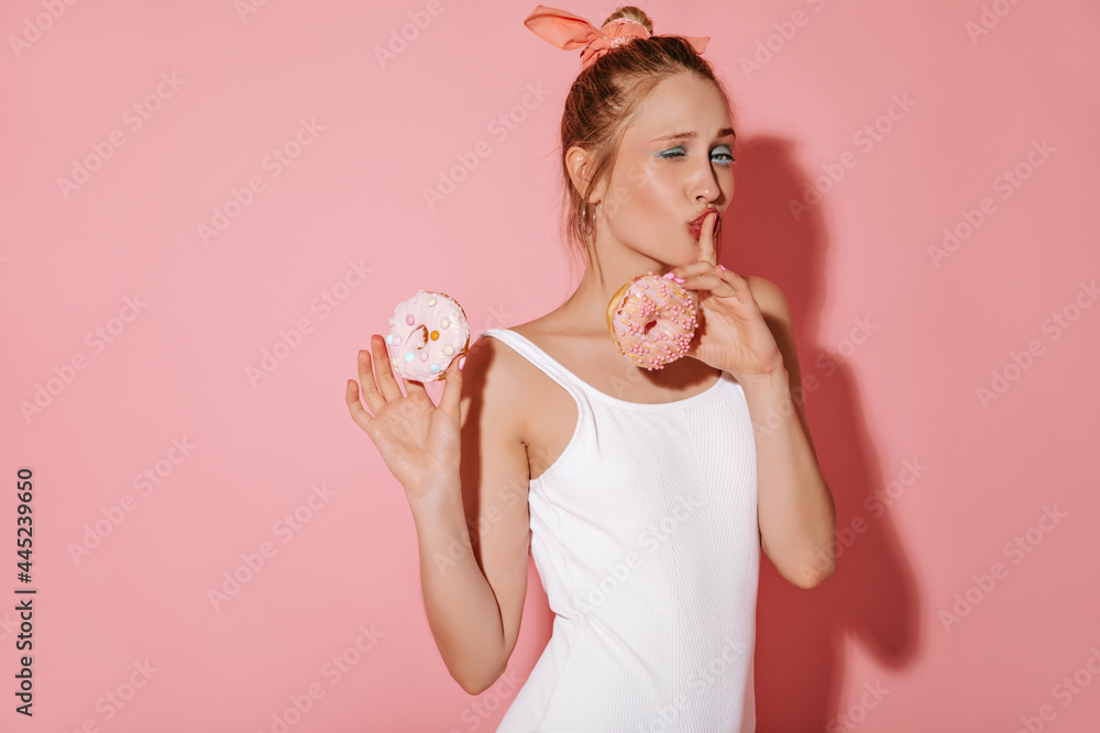 Modern woman with blonde hair and stylish makeup in white cool swimsuit posing with two donuts and winking on isolated backdrop.. Modern woman with blonde hair and stylish makeup in white cool swimsuit posing with two donuts and winking on isolated backdrop..