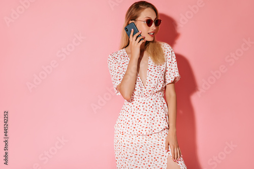 Stylish woman with blonde hairstyle in modern red sunglasses and trendy light dress talking on phone on pink background..