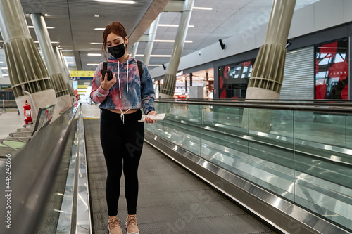 latina tourist woman with mask moving along the airport treadmill with social distancing while using her smart phone during the pandemic of coronavirus or covid19 virus, concept of new normality photo