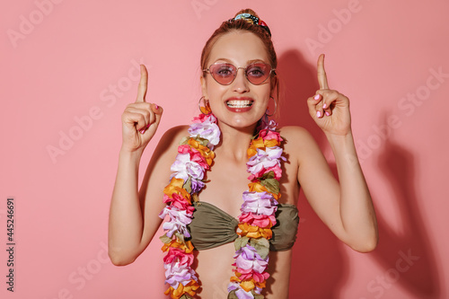 Cheerful girl with pink sunglasses in cool khaki swimsuit and bright necklace of flowers smiling and pointing to place for text on isolated backdrop..