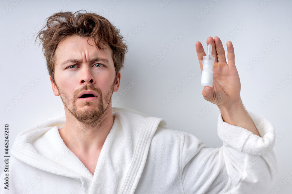 Closeup of young man in bathrobe with sickness using nasal spray for congested nose. Sick man using nose drops for runny nose. Coronavirus COVID-19 symptoms. Isolated white background, copy space
