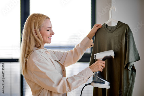 Blonde woman using steaming iron to ironing fashion shirt at home in bright cozy room. Female holding electric steamer, using modern technologies. Copy space. Side view