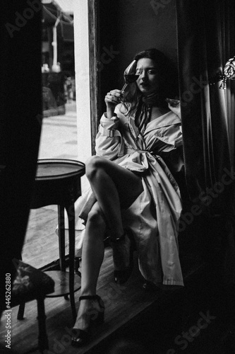 Elegant lady resting in bar with wine glass. Black and white portrait of young beautiful woman dressed in trench coat