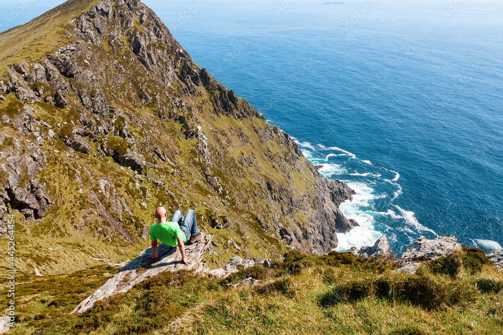 Bald male tourist in green shirt on the edge of a rock. Achill island, county Mayo, Ireland. Warm sunny day, blue sky and water.