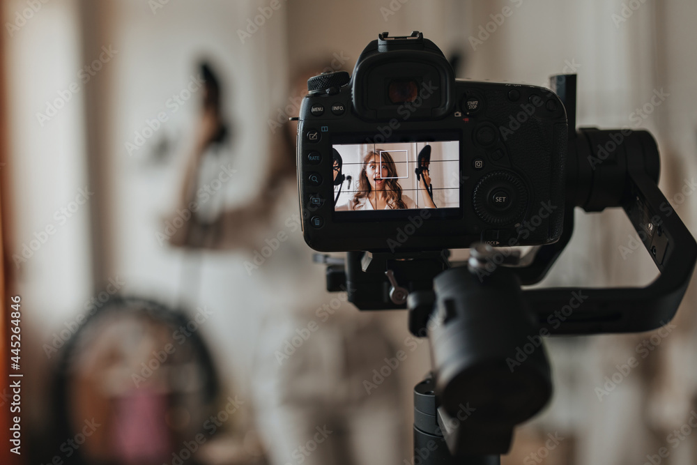 Snaphot of camera taking photo of curly woman. Brunette girl holding black shoes and posing in white cozy room