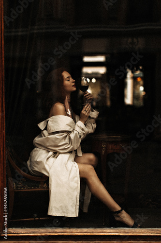 Woman in beige trench drinking wine. Attractive young lady in stylish outfit resting in cozy cafe and enjoying champagne