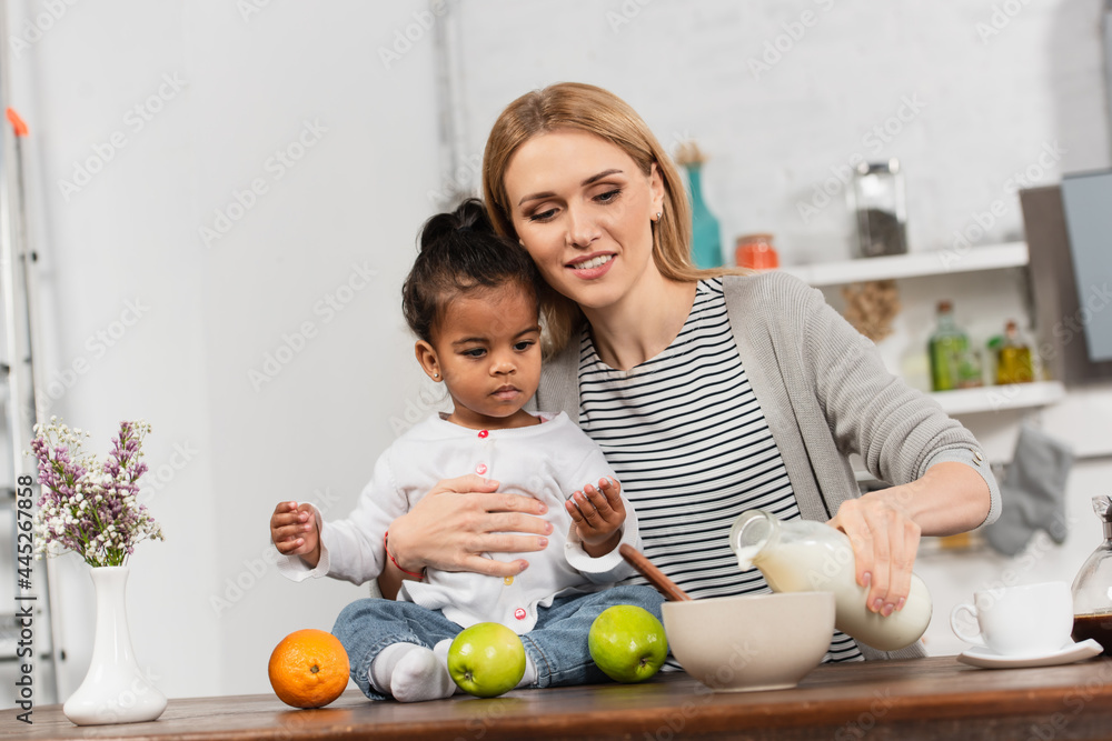 Fototapeta premium cheerful woman pouring milk in bowl and holding adopted african american daughter