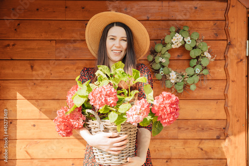 A young horticulturist holds a red hydrangea and smiles. Woman with a straw hat is standing in the garden with a wooden garden shed in the background. Gardening with fun. Life in the countryside
