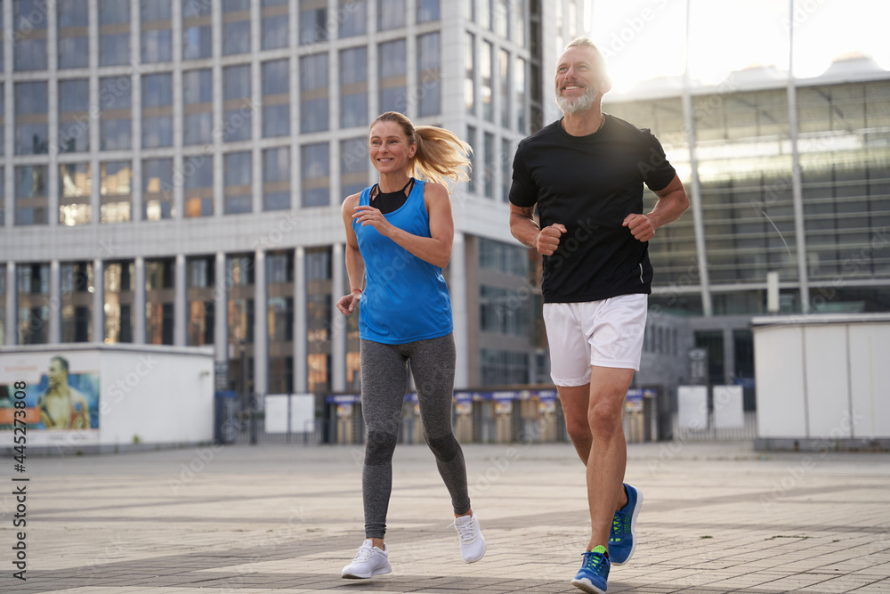 Full length shot of motivated middle aged couple, man and woman in sportswear running together in the city, having morning workout