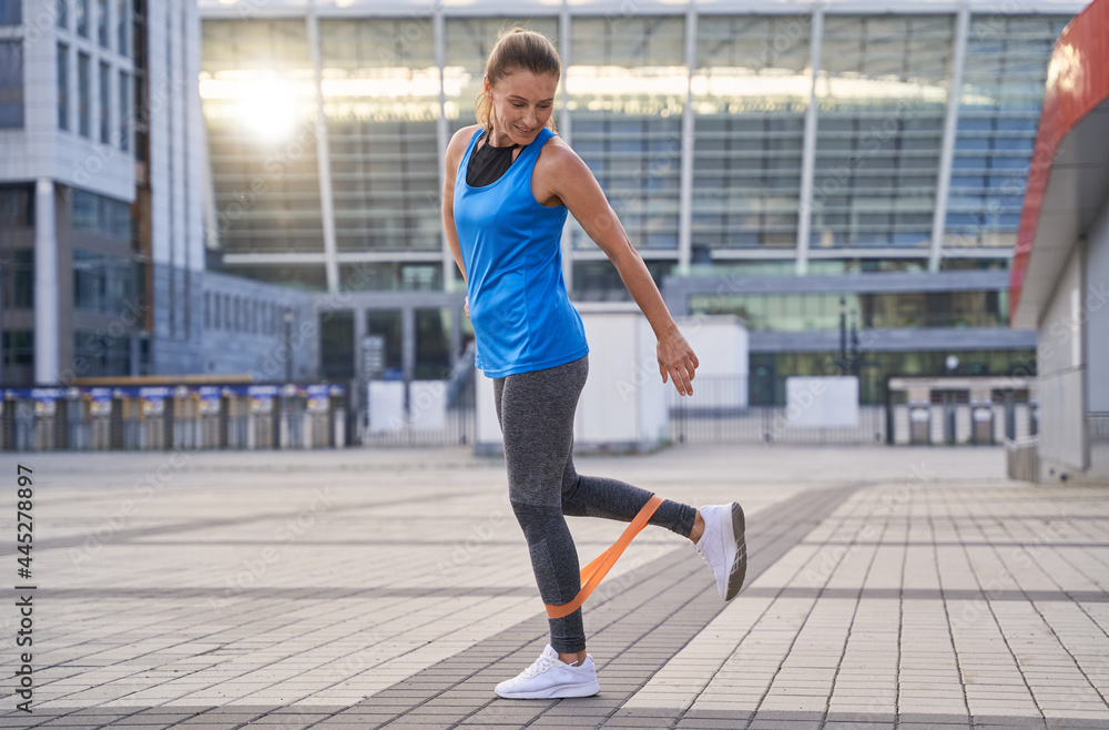 Full length shot of active sporty woman in sportswear warming up using resistance band while standing outdoors, exercising in the city