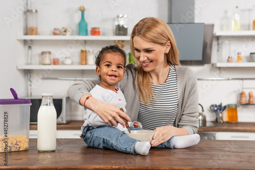 happy adopted african american kid holding spoon near bowl while sitting on kitchen table near smiling mother