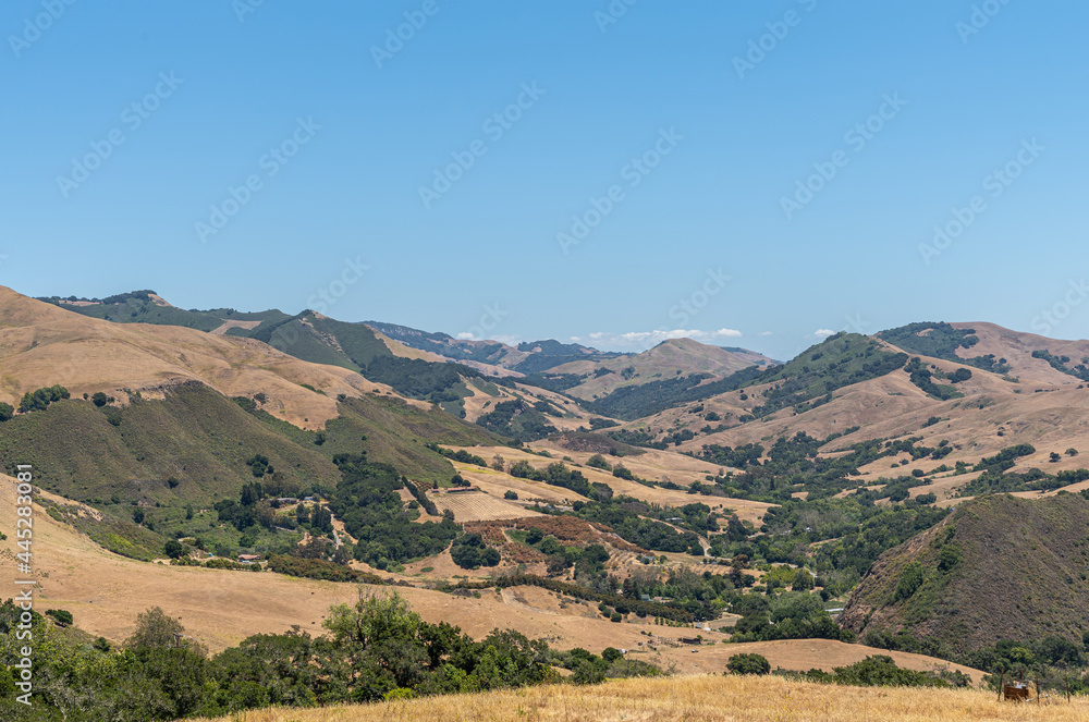 Cambria, CA, USA - June 9, 2021: Wide landscape of Back country with dry ranch land and patches of green trees on steep flanks of mountains under blue sky.