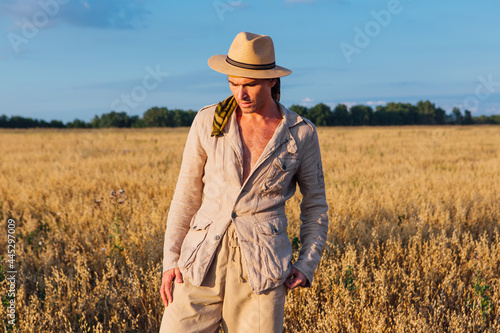 Tall handsome man dressed in a coarse linen suit and hat standing at golden oat field