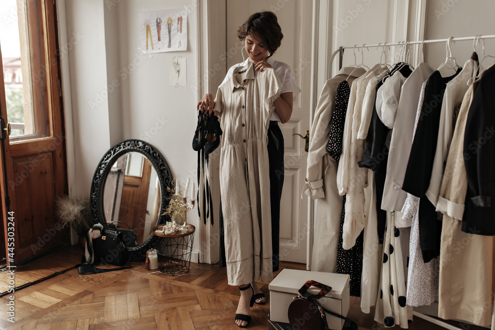 Attractive dark-haired young woman in midi skirt, white blouse smiles, holds stylish linen beige dress and black shoes, poses in cozy room. Attractive dark-haired young woman in midi skirt, white blouse smiles, holds stylish linen beige dress and black shoes, poses in cozy room.