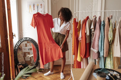 Fotografiet Curly stylish dark-skinned woman in khaki shorts and white blouse holds hanger with long red dress and poses in dressing room