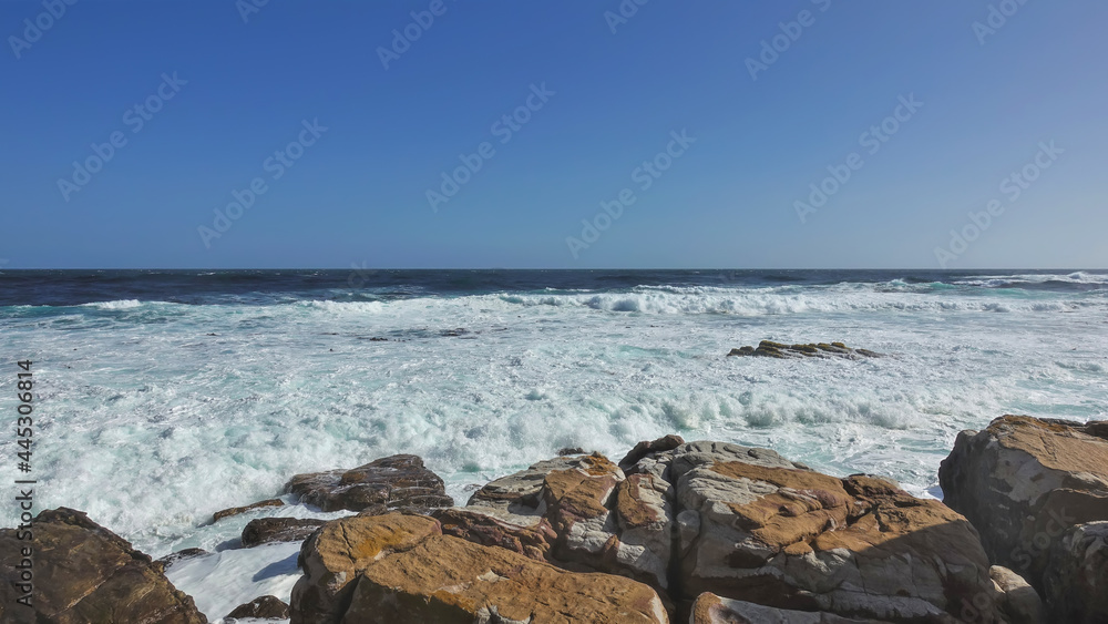 Cape of Good Hope in Cape Town. The waves of the turquoise Atlantic Ocean break on the coastal boulders and a dense white foam is formed. Clear blue sky 