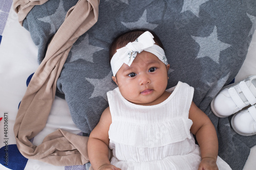 Healthy adorable baby in white dress lying down on bed, eyes looking away. Top view portrait of a southeast asian race baby girl.