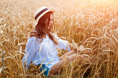Happy girl sits in wheat field in straw hat. Cute young woman of Caucasian ethnicity in casual clothes enjoys ripe golden wheat in a field. Free woman concept