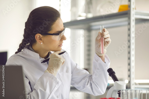 Thoughtful female scientist wearing protective eye glasses looking at medical test tube glass flask working do research in scientific laboratory closeup side view portrait. Pharmacology and medicine